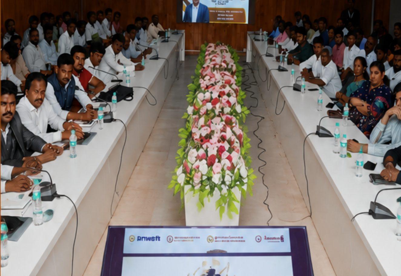 A full district meeting hall in Ramanathapuram, with attendees seated along both sides of a long table sharing a few microphones